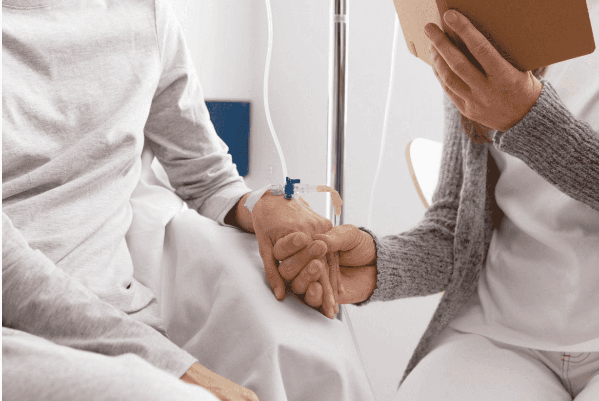 A person holding the hand of a patient with an IV while reading a book.
