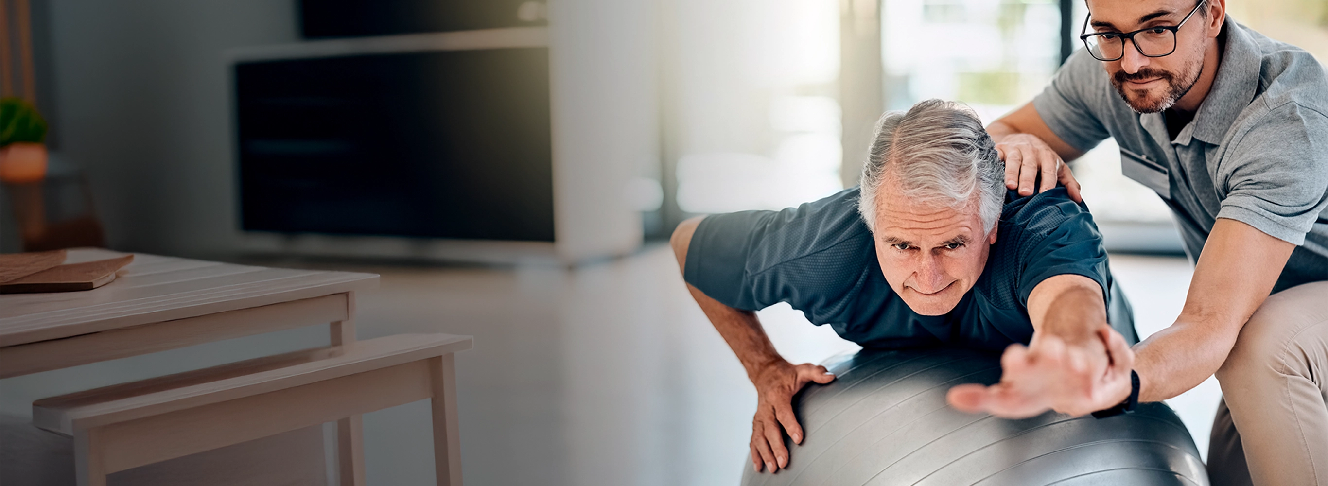 A senior man and his physical therapist doing balance exercises with a large stability ball in a sunlit living room.