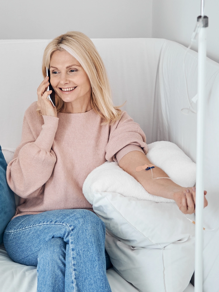 A healthcare professional using a stethoscope to examine a smiling female patient in her living room, with a clinical IV hydration setup positioned nearby.