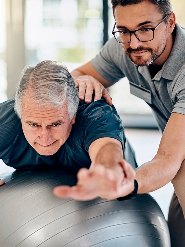A senior man and his physical therapist doing balance exercises with a large stability ball in a sunlit living room.