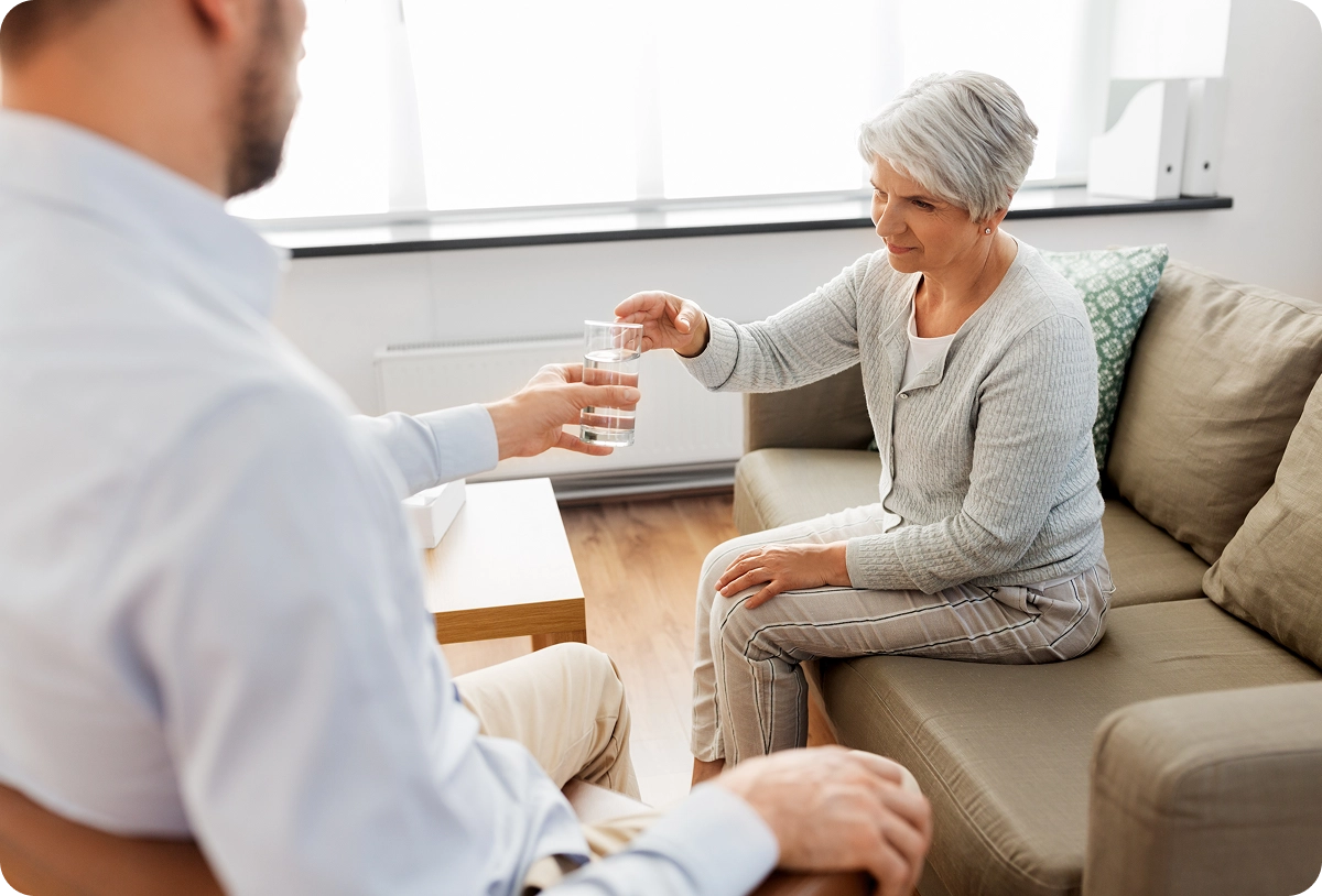 A healthcare professional or therapist gently handing a bottle of medication or comfort item to an elderly woman, possibly a cancer patient, sitting on a couch.